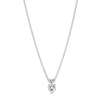 A silver necklace with two heart-shaped pendants, one larger than the other, both adorned with clear stones. The necklace is displayed against a plain white background.