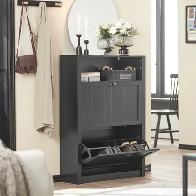 A dark gray shoe cabinet with two flip-down doors is shown in a hallway or entryway. A coat and a purse hang on a rack to the left, and a mirror and decorative items are on top of the cabinet.