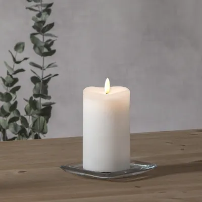 A white LED candle with a flickering flame sits on a square glass coaster on a wooden table. In the background, a grey wall is visible with a branch of eucalyptus leaves.