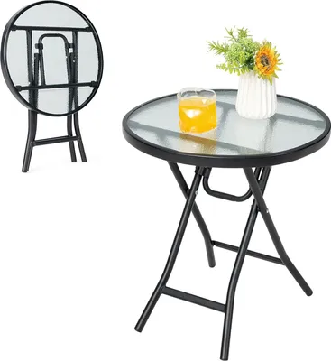 A round glass-top folding table with a metal frame, shown with a glass of orange juice and a small vase of flowers.