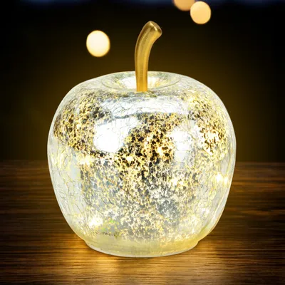 A decorative glass apple with LED lights sits on a wooden surface, with blurred lights in the background.