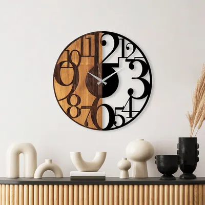 A modern wall clock with a split design, half wood and half black metal numbers, displayed on a white wall above a wooden cabinet with decorative vases.