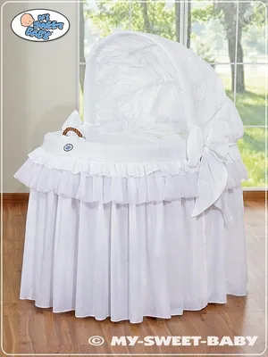 A white baby bassinet with a canopy and ruffled skirt, placed indoors near a window with a view of trees.