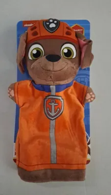 A plush hand puppet of the Paw Patrol character Zuma is shown against a light gray background, with its packaging visible behind it.