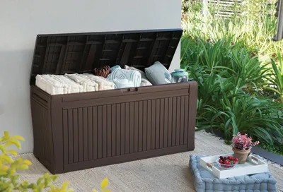An outdoor storage box filled with cushions and blankets, placed on a patio next to lush greenery.