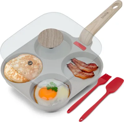 A multi-compartment frying pan with a glass lid, containing a pancake, bacon, and a fried egg. Also includes red silicone cooking utensils.