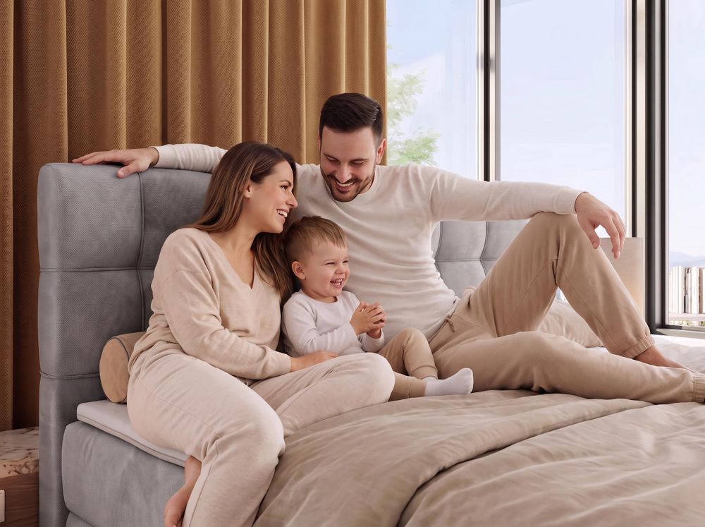 Familie sitzt gemeinsam auf einem grauen Polsterbett mit beiger Bettwäsche.