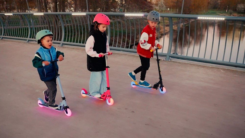 Drie kinderen rijden samen op steps met helmen op een brug.