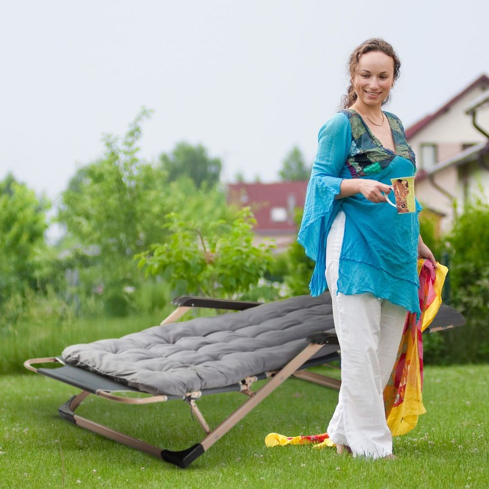 Frau mit Tasse und Tuch neben grauer Gartenliege im Garten.