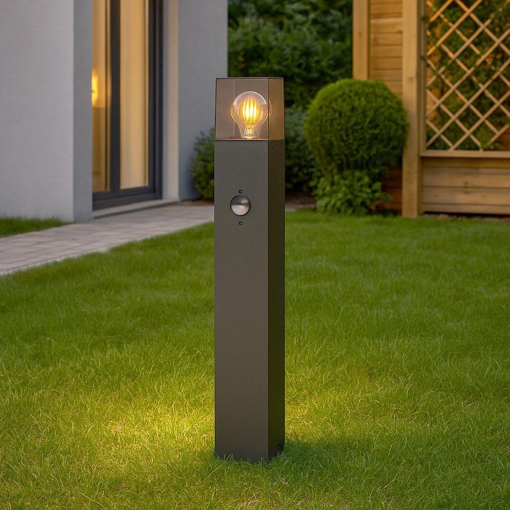 Schwarze Außenleuchte mit Bewegungsmelder und Filamentlampe im Garten neben Haus und Holzpavillon.