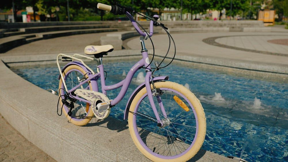 Bicicleta infantil lavanda junto a una fuente, mostrando su diseño y accesorios.