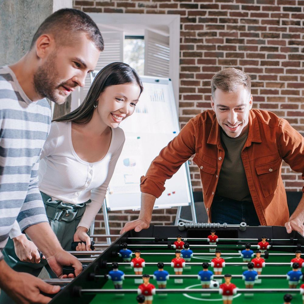 Drei Erwachsene (zwei Männer, eine Frau) spielen lächelnd gemeinsam Tischfußball