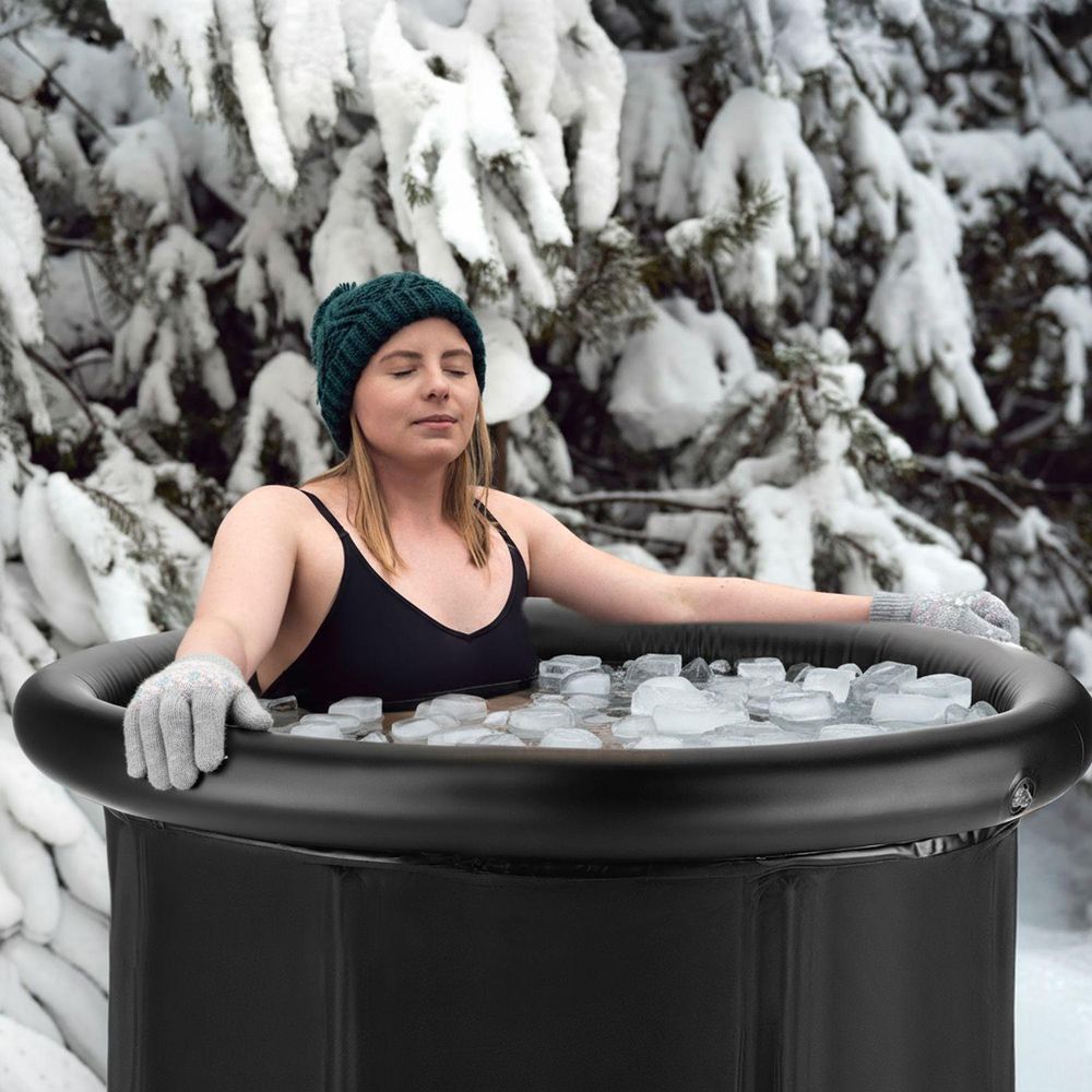 Frau in schwarzem Bikini und Mütze genießt ein Eisbad in der kedica Tonne vor verschneiten Bäumen