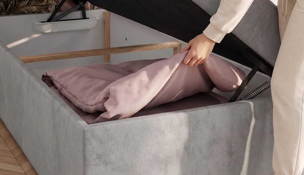 Graues Polsterbett mit geöffnetem Bettkasten und rosa Decke im Stauraum.