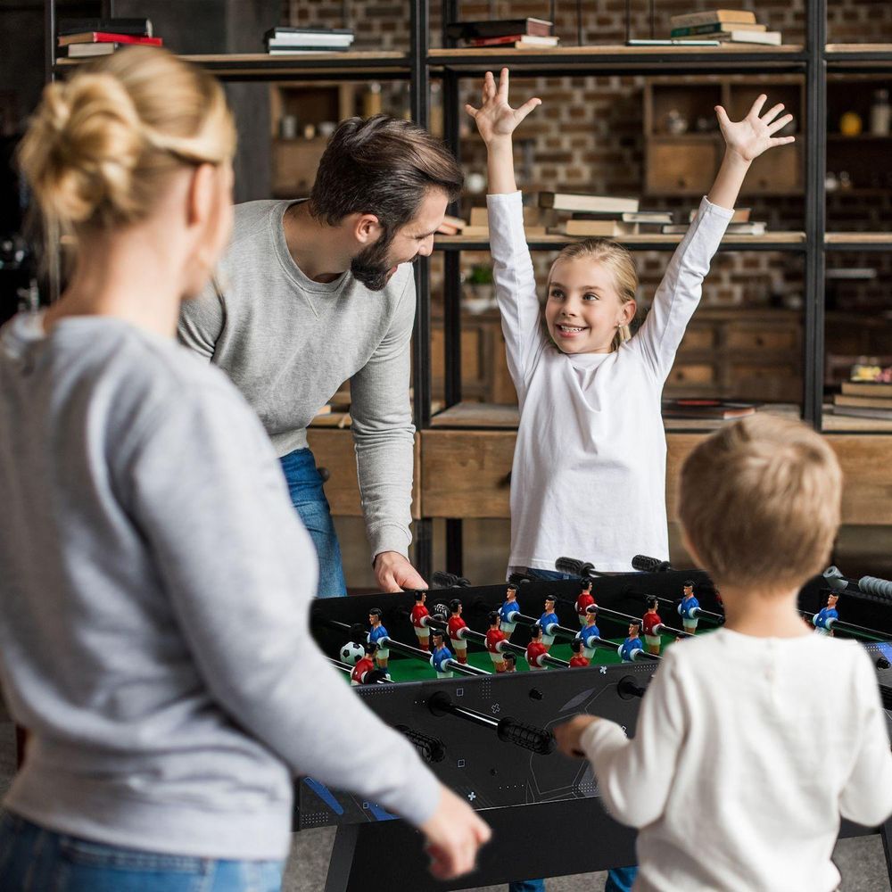 Familie (Mutter, Vater, zwei Kinder) spielt Kicker; das Mädchen jubelt mit erhobenen Händen