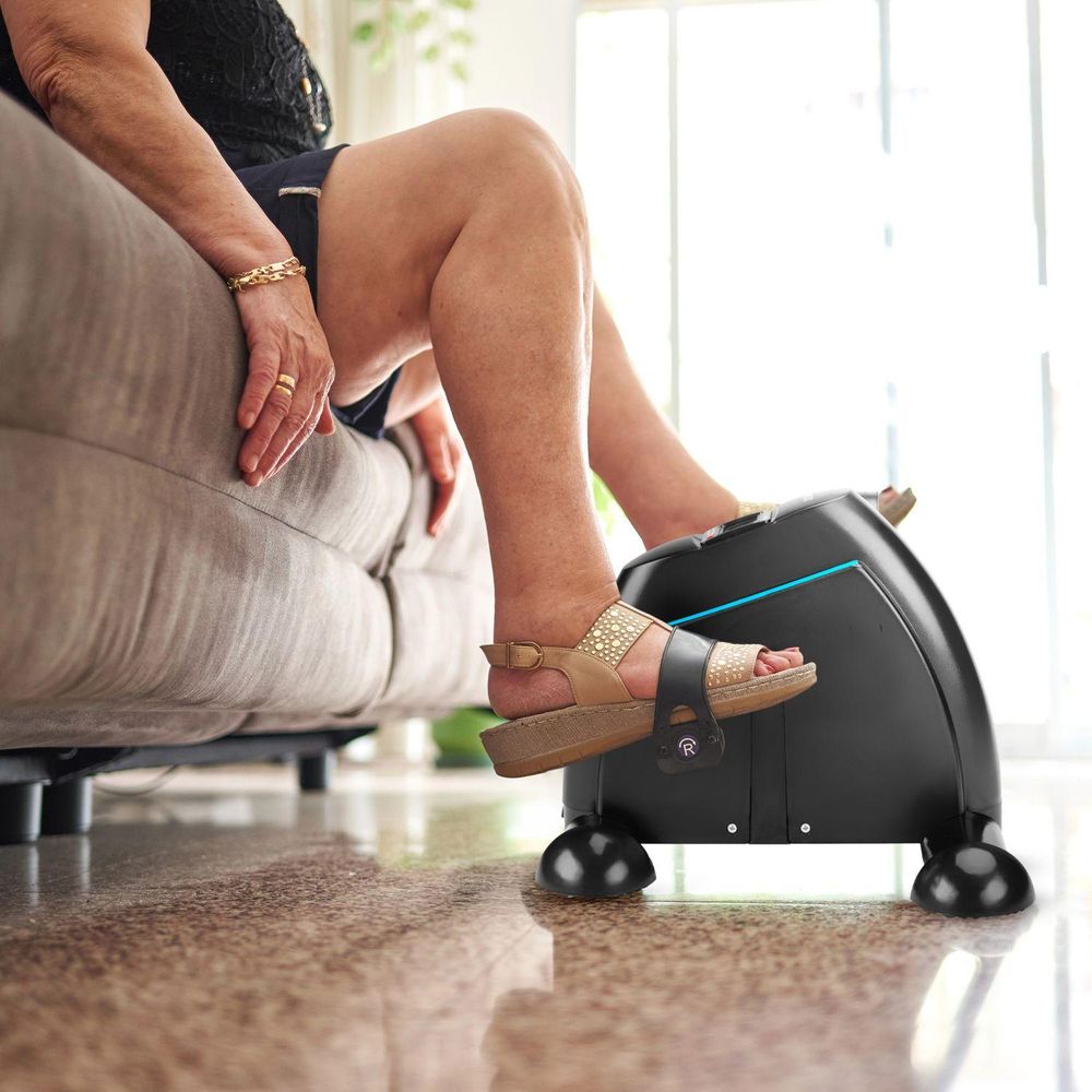Nahaufnahme: Fuß in Sandale auf Pedal des Mini-Heimtrainers. Frau sitzt auf Couch