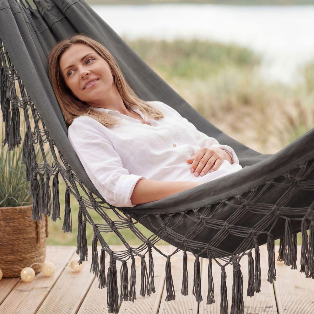 Frau entspannt lächelnd in einer grauen Hängematte mit Fransen auf einer Holzterrasse