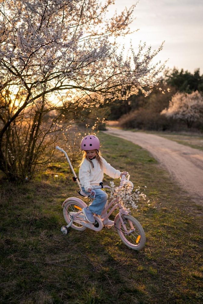 Niña montando la bicicleta rosa al aire libre con casco y barra de empuje.