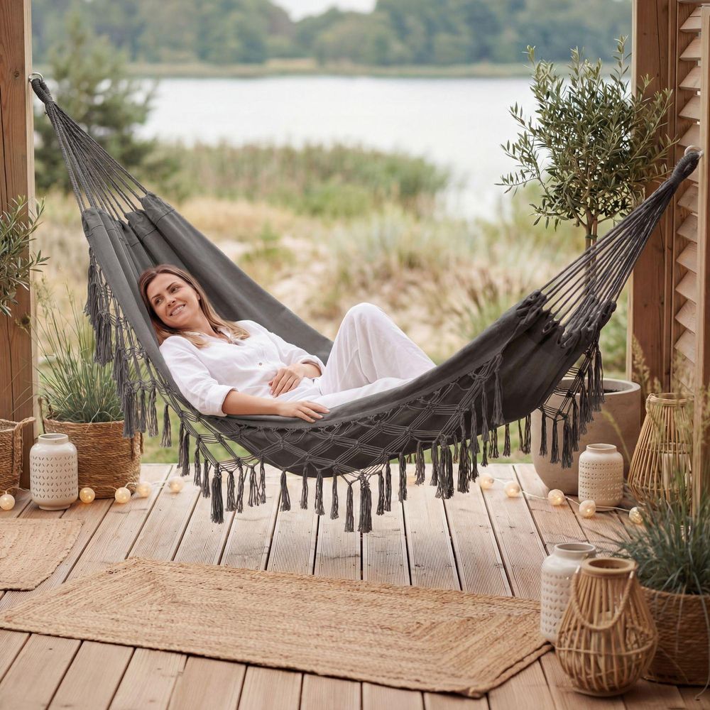 Frau liegt in grauer Boho-Hängematte auf Terrasse am See, umgeben von Deko und Pflanzen