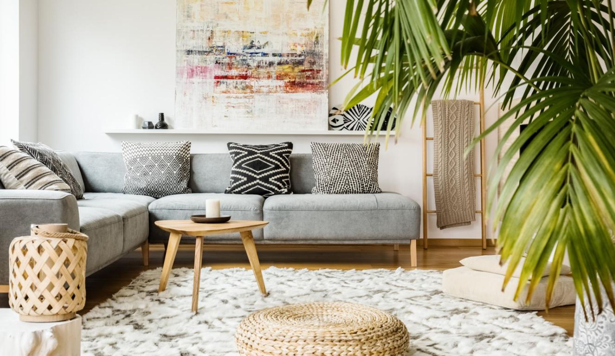Wohnzimmer mit grauer Couch, Teppich, großer Pflanze, Beistelltisch, Sitzpuff und mit einem Bild.