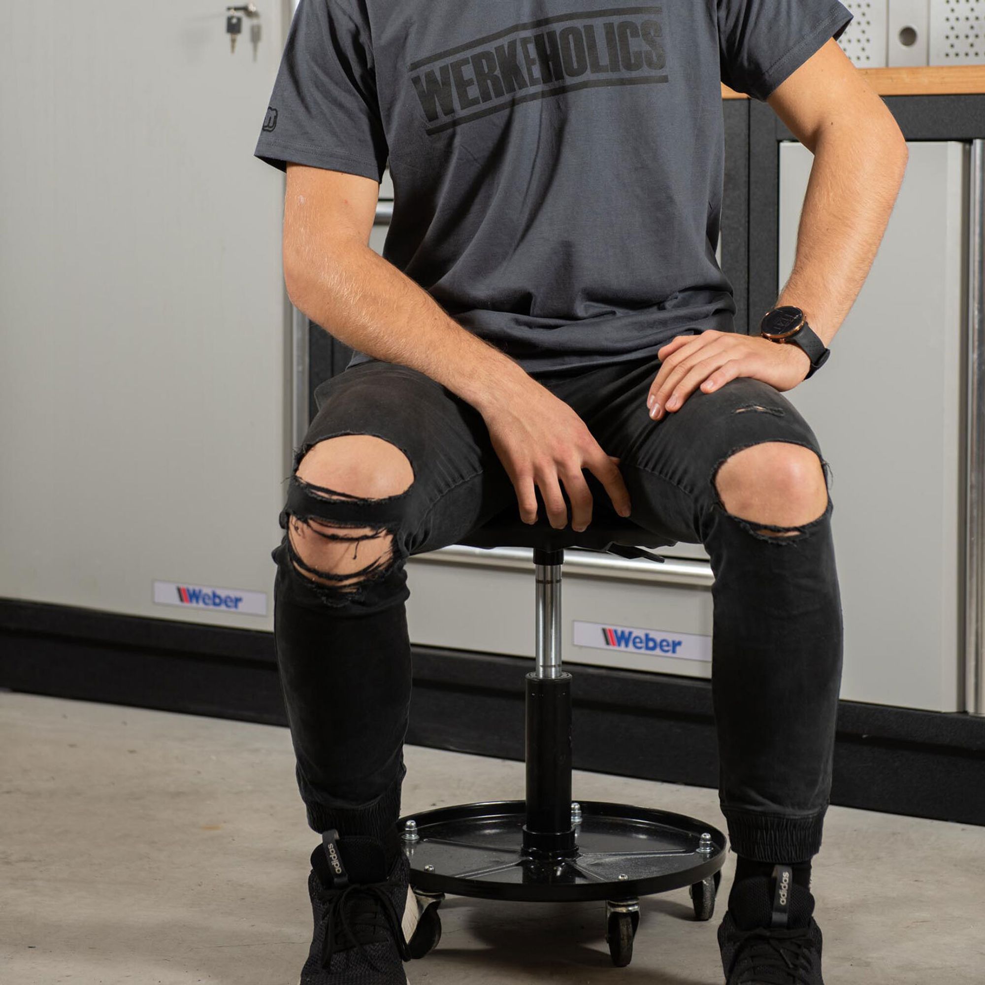 Das Bild zeigt eine auf dem Hocker sitzende Person. Im Hintergrund ist eine Werkstattwand zusehen.