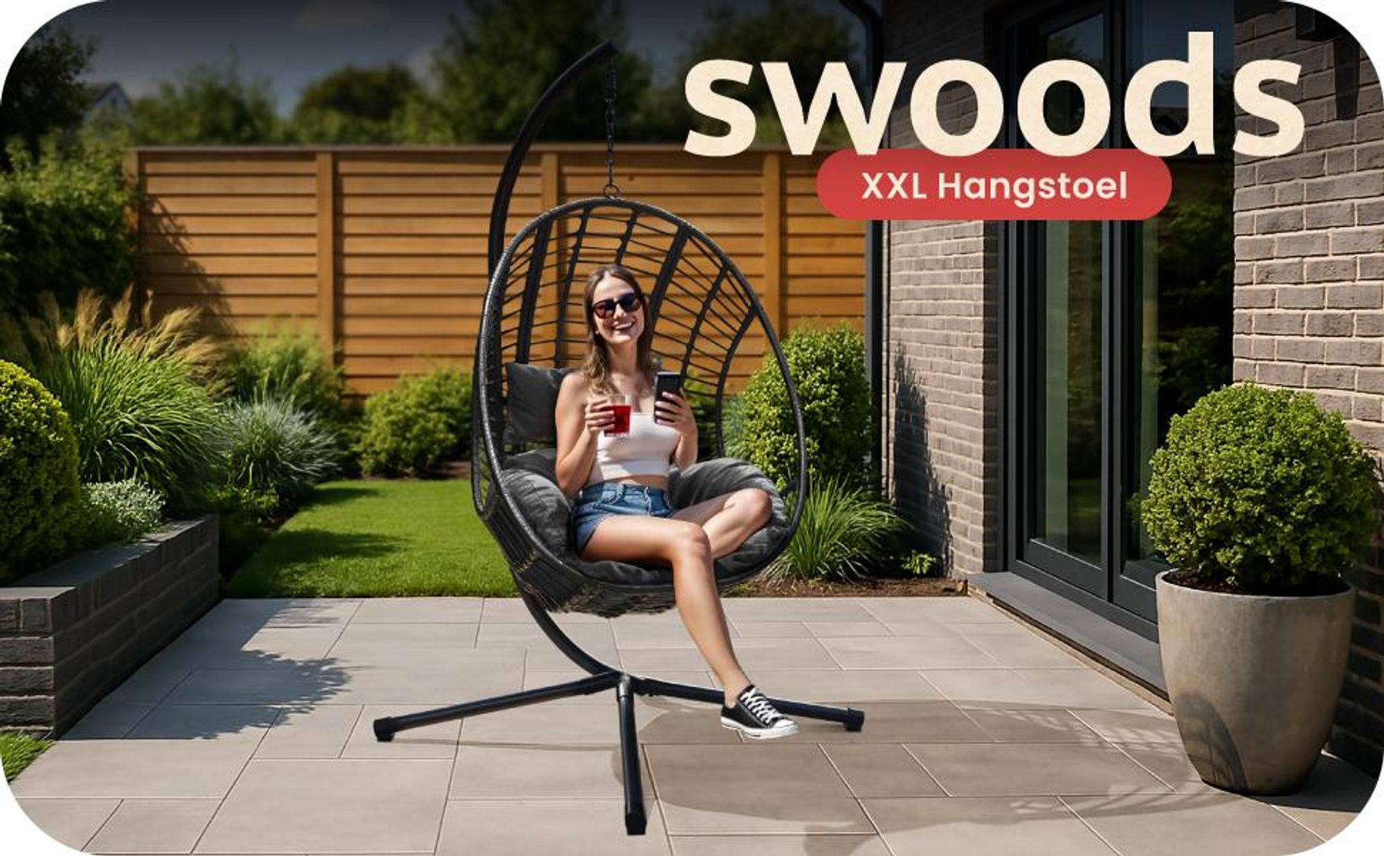 Duże czarne krzesło wiszące w kształcie jajka z poduszką na patio. Kobieta relaksująca się na krześle z napojem. Widoczny tekst „Swoods XXL Hangstoel”. Zewnętrzna część ogrodu i domu w tle.