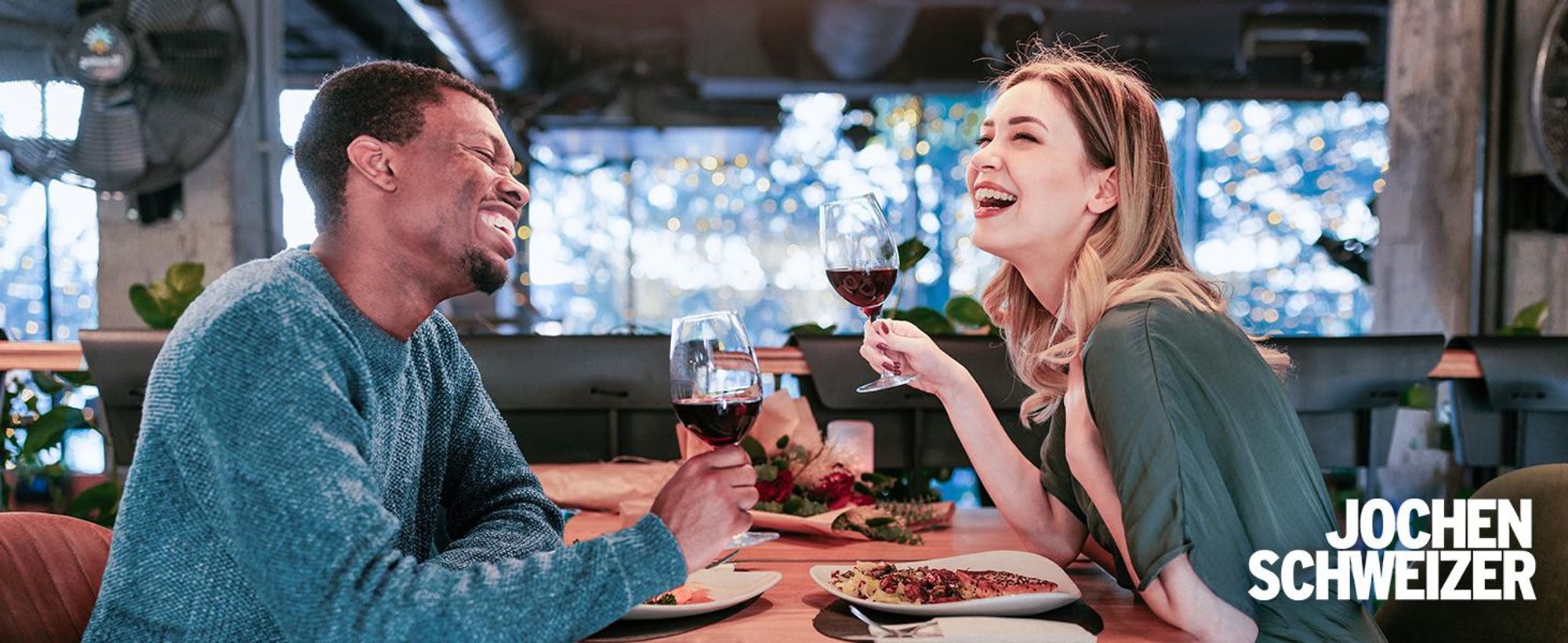 Das Bild zeigt ein Paar bei einem Dinner. Beide lachen und halten ein Glas Wein in der Hand.